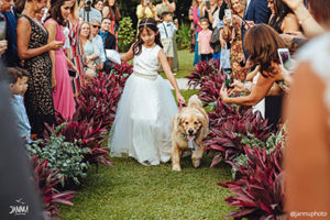 Young flower girl in white dress walking down a wedding aisle with a golden retriever dog.