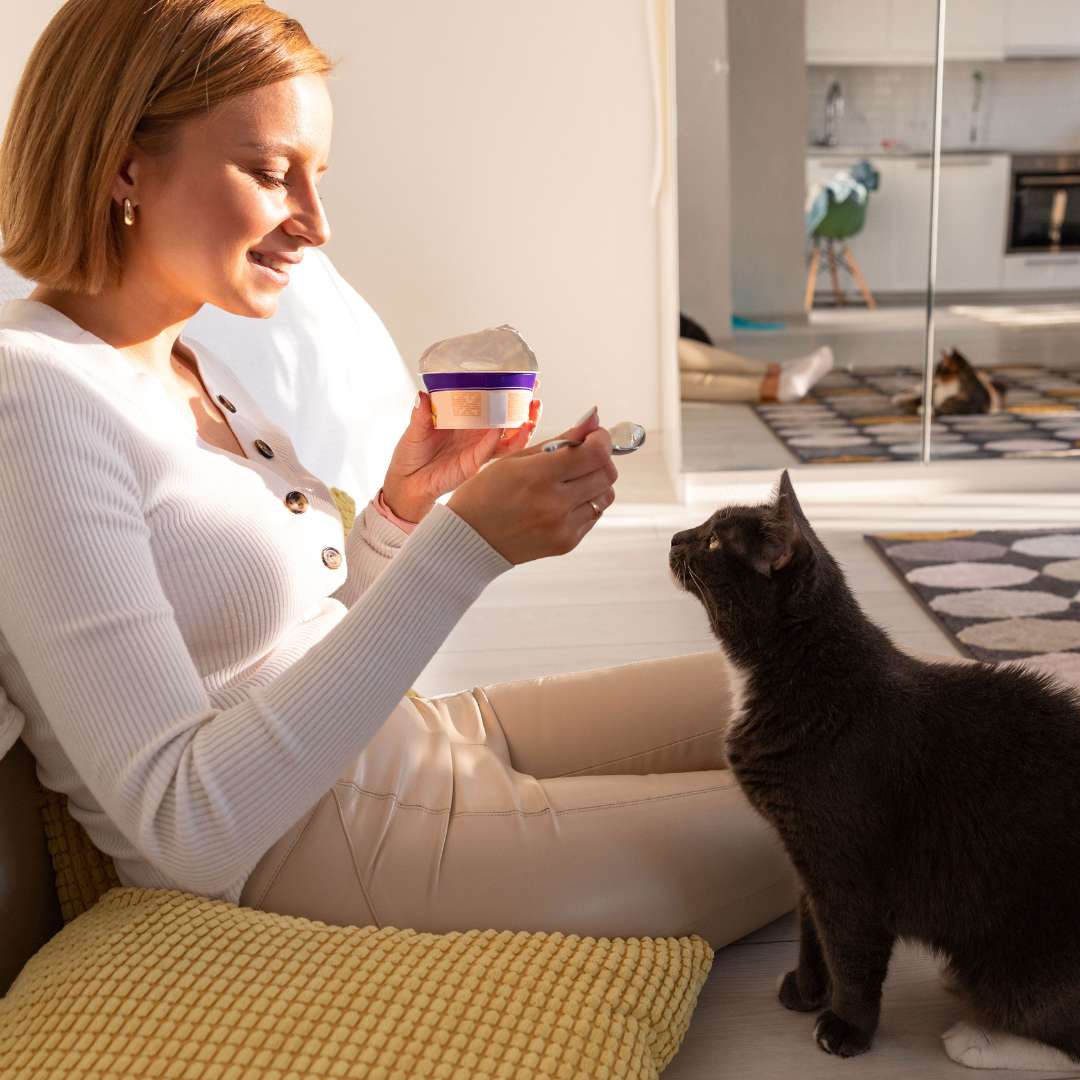 A woman in a white, long-sleeved buttoned-up sweater feeds her black and white cat unsweetened yogurt because he or she has cat and dog upset tummy. 