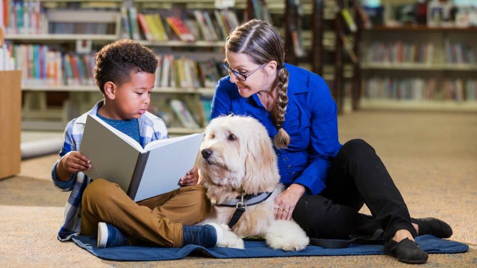 A dog in a library reading with a child and woman.