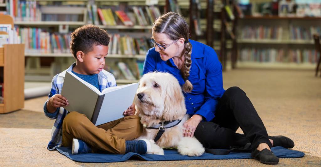 A dog in a library reading with a child and woman.