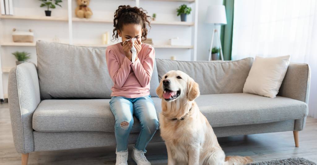 A sick girl sits on a couch with a dog nearby.